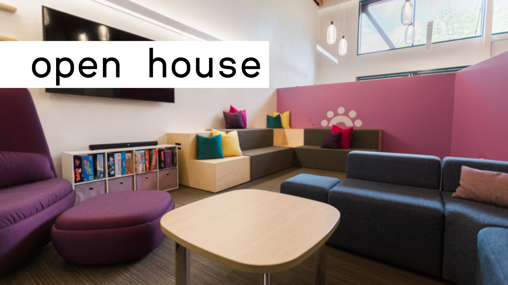 Colorful, open, and airy recreational room with furniture, shelves with board games, and a television, as well as windows. The top left corner of the image includes black text which reads "open house" in front of a white background.